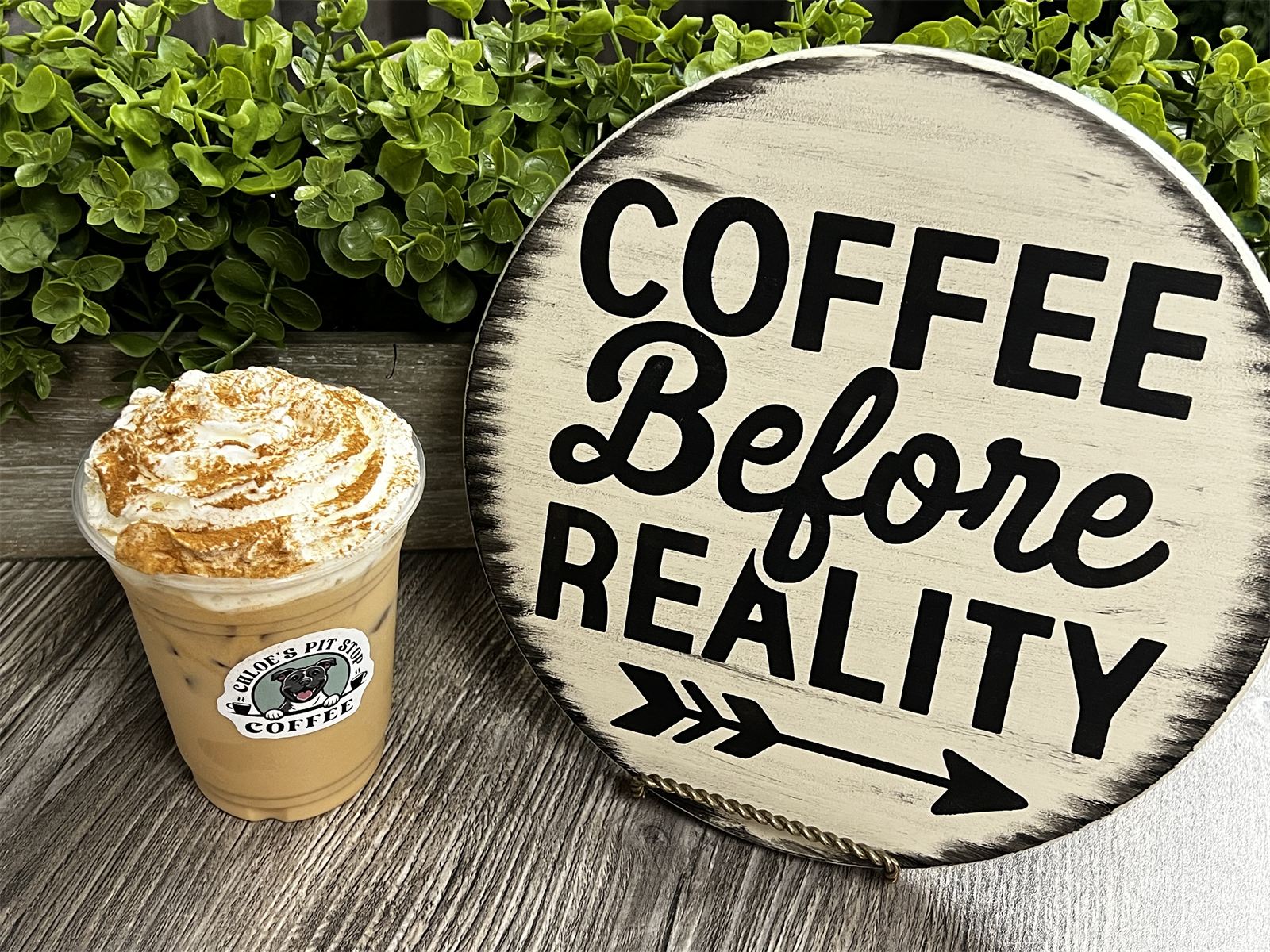 An iced latte topped with whipped cream and cinnamon, wearing a Chloe's Pit Stop sticker, next to a wooden Coffee Before Reality sign