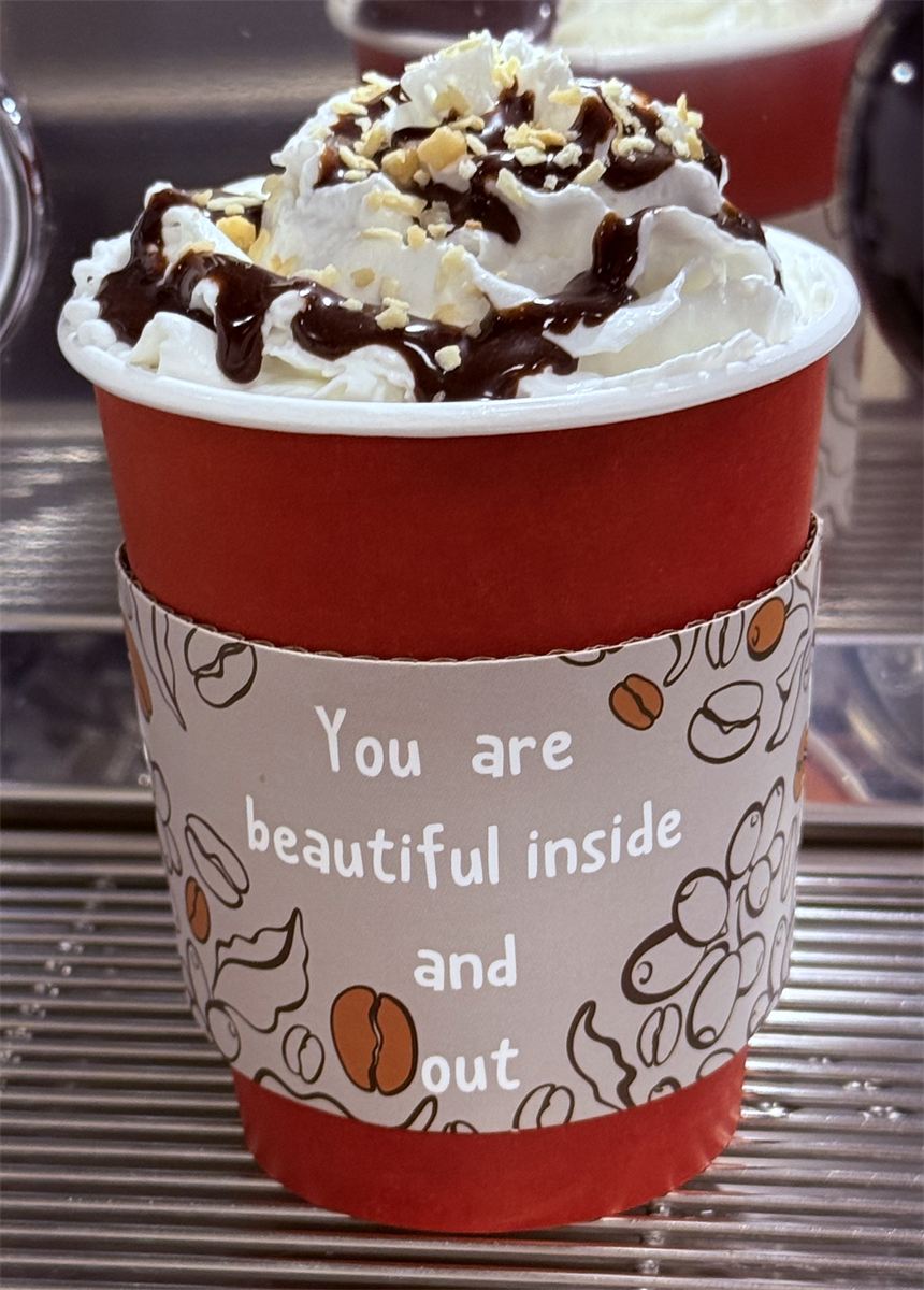 A hot drink in a red cup piled with whipped cream, chocolate drizzle, and chopped nuts, sleeve reading you are beautiful inside and out